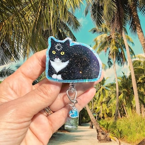 May include: A black and white cat-shaped badge reel with a glittery finish, yellow eyes, and a loading icon on its forehead. The badge reel features a clear plastic strap and a small, dangling blue heart charm attached to the metal hardware.