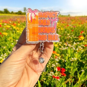 Pu&ograve; includere: Un portabadge glitterato con la scritta "SUSPECT ONLY DRINKS Red Bull". Il badge presenta un design di lattina Red Bull arancione con fiocchi rosa. Il portabadge ha una base in plastica trasparente e un bottone argentato.