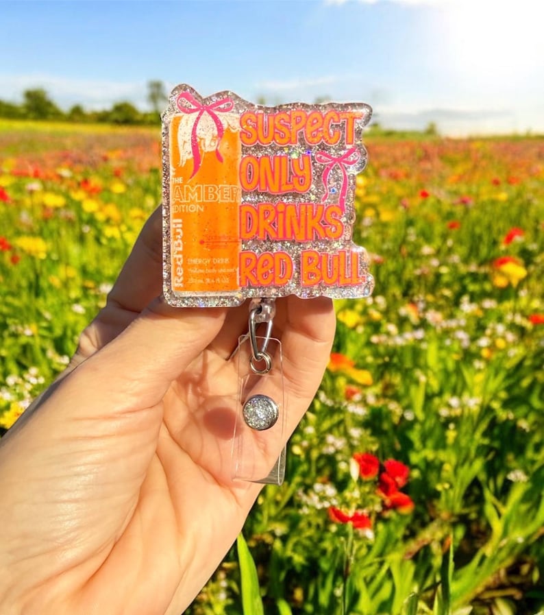 Pu&ograve; includere: Un portabadge glitterato con la scritta "SUSPECT ONLY DRINKS Red Bull". Il badge presenta un design di lattina Red Bull arancione con fiocchi rosa. Il portabadge ha una base in plastica trasparente e un bottone argentato.