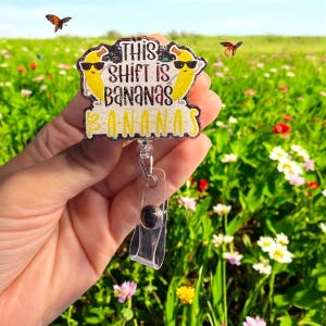 May include: A badge reel with the text "THIS SHIFT IS BANANAS" and two cartoon bananas wearing sunglasses. The badge reel is yellow, black, and white, with a clear plastic clip and a black button. Butterflies and flowers are in the background.