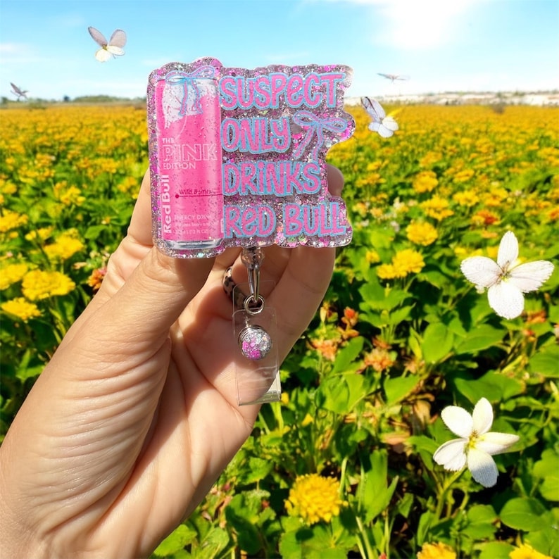 Pu&ograve; includere: Un portabadge rosa glitterato con la scritta "Suspect Only Drinks Red Bull". Il design include una lattina di Red Bull rosa. Lo sfondo mostra un campo di fiori gialli e un cielo blu.