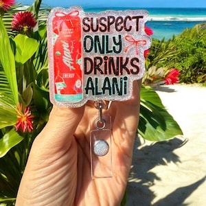 Pu&ograve; includere: Un porta badge trasparente con un bordo glitterato e la scritta "SUSPECT ONLY DRINKS ALANI". Il badge presenta una grafica di lattina di bevanda energetica Alani Nu rossa. Il porta badge &egrave; tenuto in mano su uno sfondo di spiaggia.