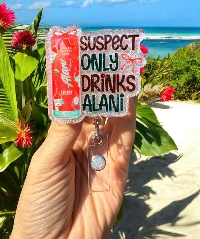 Pu&ograve; includere: Un porta badge trasparente con un bordo glitterato e la scritta "SUSPECT ONLY DRINKS ALANI". Il badge presenta una grafica di lattina di bevanda energetica Alani Nu rossa. Il porta badge &egrave; tenuto in mano su uno sfondo di spiaggia.
