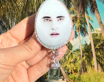 May include: An oval-shaped badge reel featuring a glittery white surface with a printed image of a person's face. The badge reel has a silver-toned glitter border and a clear plastic strap with a small silver heart-shaped charm attached.