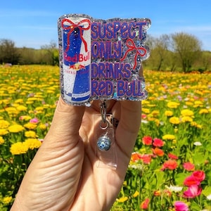Pu&ograve; includere: Un portabadge glitterato con un design a lattina Red Bull e la scritta "SUSPECT ONLY DRINKS RED BULL" in blu e rosso. Il portabadge ha un supporto in plastica trasparente e un cordino retrattile.