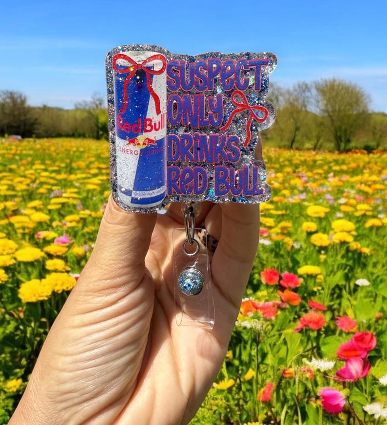 Pu&ograve; includere: Un portabadge glitterato con un design a lattina Red Bull e la scritta "SUSPECT ONLY DRINKS RED BULL" in blu e rosso. Il portabadge ha un supporto in plastica trasparente e un cordino retrattile.