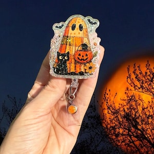 May include: A Halloween-themed badge reel featuring a ghost, black cat, and jack-o'-lantern. The design is set against a dark background with an orange moon. The reel has a clear plastic body and an orange accent.