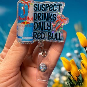 Pu&ograve; includere: Un portabadge glitterato con un design a lattina di Red Bull e la scritta "SUSPECT DRINKS ONLY RED BULL". Il portabadge ha un cordino retrattile in plastica trasparente e un bottone decorativo.
