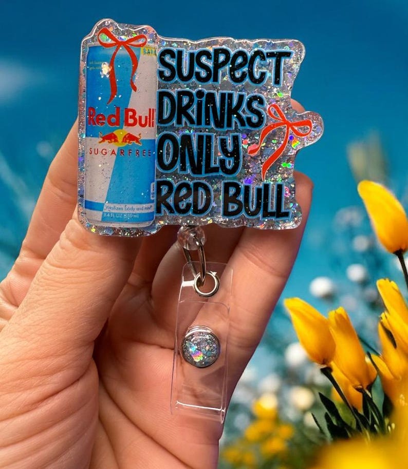 Pu&ograve; includere: Un portabadge glitterato con un design a lattina di Red Bull e la scritta "SUSPECT DRINKS ONLY RED BULL". Il portabadge ha un cordino retrattile in plastica trasparente e un bottone decorativo.