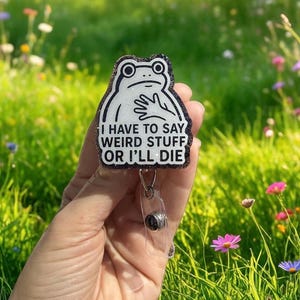 May include: A white frog-shaped badge reel with a black glitter border. The frog has a sad expression and a hand on its chest. The text on the badge reel reads "I HAVE TO SAY WEIRD STUFF OR I'LL DIE."