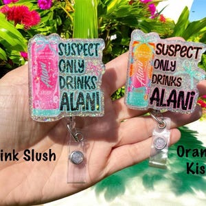 Pu&ograve; includere: Due porta badge con la scritta "SUSPECT ONLY DRINKS ALANI". Uno presenta un design di lattina rosa con l'etichetta "Pink Slush", l'altro ha un design di lattina arancione con l'etichetta "Orange Kiss". Entrambi sono tenuti in mano.