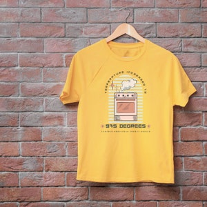 Op de afbeelding: Geel T-shirt met een retro-stijl illustratie van een oven met de tekst "Temperatuur verhoogd naar 375 graden" en "Nog een kerstkalkoen geroosterd".