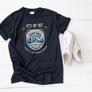 May include: Navy blue t-shirt with a graphic featuring a Bigfoot illustration and the text "Yeti or Not." The design includes the words "Spirit of Exploration" and "Legend of the Forbidden Mountain." White canvas sneakers are next to the shirt.