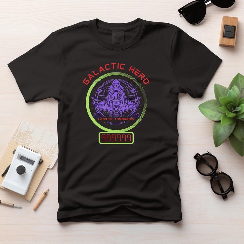 K&ouml;nnte beinhalten: Schwarzes T-Shirt mit einem Grafikdesign, das ein lila Raumschiff in einem gr&uuml;nen Kreis zeigt, die Worte "GALACTIC HERO" in Rot und "LAND OF TOMORROW" darunter. Eine Digitalanzeige zeigt "999999".