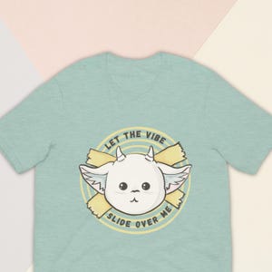 May include: A light blue t-shirt featuring a cartoon goat illustration with the text "LET THE VIBE SLIDE OVER ME" in a circular design. The goat has white fur, black eyes, and small horns. The shirt is made of soft material.