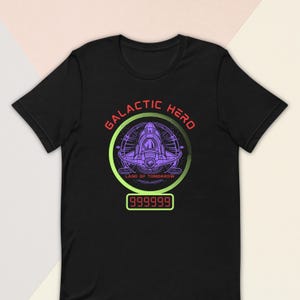 May include: Black t-shirt with a graphic design featuring a purple spaceship inside a green and purple circle. The text "GALACTIC HERO" is in red, and "LAND OF TOMORROW" is in green. A digital display shows "999999".