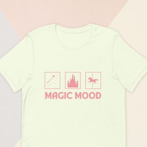 May include: A light green t-shirt with a pink graphic that includes a wand, a castle, and a carousel horse. The words "MAGIC MOOD" are printed below the graphics in pink block letters.