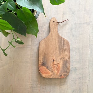 May include: A wooden cutting board with a brown leather handle. The board has a natural wood grain and a light brown stain.