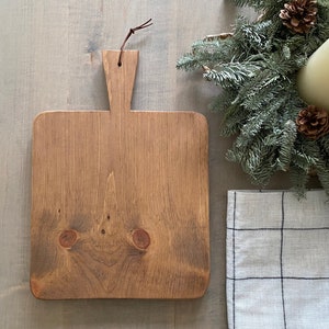 May include: A square wooden cutting board with a brown stain and a leather handle. The board is sitting on a light wood surface.
