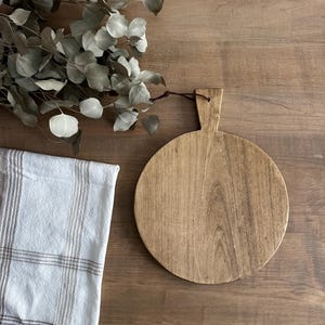 Op de afbeelding: Een ronde houten snijplank met een handvat, weergegeven op een houten oppervlak. Een takje eucalyptus en een wit en grijs gestreepte handdoek zijn ook aanwezig. De snijplank is lichtbruin van kleur.