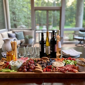 May include: A large wooden serving board filled with a variety of snacks, including crackers, cheese, fruit, vegetables, and meat. There are also several bottles of wine and glasses on the table.