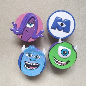 May include: Four round, painted wooden decorations featuring characters from the animated film "Monsters, Inc.". The characters include Celia Mae, the "M" logo, James P. Sullivan, and Mike Wazowski. Each character is painted in bright, bold colours.