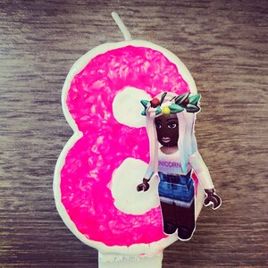 Puede incluir: Vela de cumplea&ntilde;os de color rosa n&uacute;mero ocho con un centro blanco. Una mu&ntilde;eca de ni&ntilde;a con una corona de flores, una camiseta blanca con la palabra "Unicorn" impresa en ella y una falda vaquera azul est&aacute; de pie frente al n&uacute;mero.
