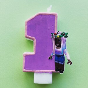 Puede incluir: Vela de cumplea&ntilde;os de color morado con el n&uacute;mero uno y una base blanca. Una ni&ntilde;a con una corona de flores y un vestido azul est&aacute; en la vela.
