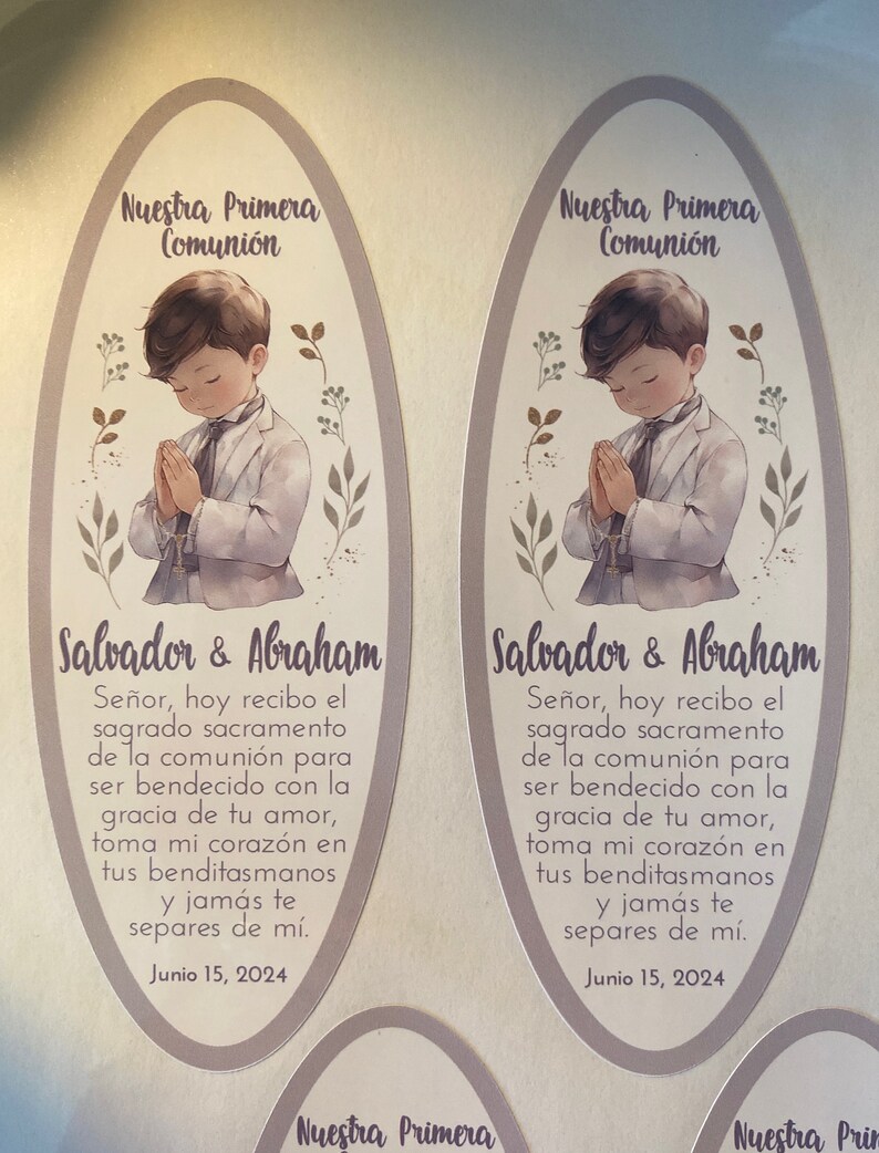 May include: Two oval-shaped cards with a light brown background and a purple border. Each card features a watercolor illustration of a young boy in a white suit praying with his hands clasped. The text "Nuestra Primera Comuni&oacute;n" is at the top of each card. The text "Salvador & Abraham" is at the bottom of each card. The text "Se&ntilde;or, hoy recibo el sagrado sacramento de la comuni&oacute;n para ser bendecido con la gracia de tu amor, toma mi coraz&oacute;n en tus benditasmanos y jam&aacute;s te separes de m&iacute;. Junio 15, 2024" is below the illustration on each card.