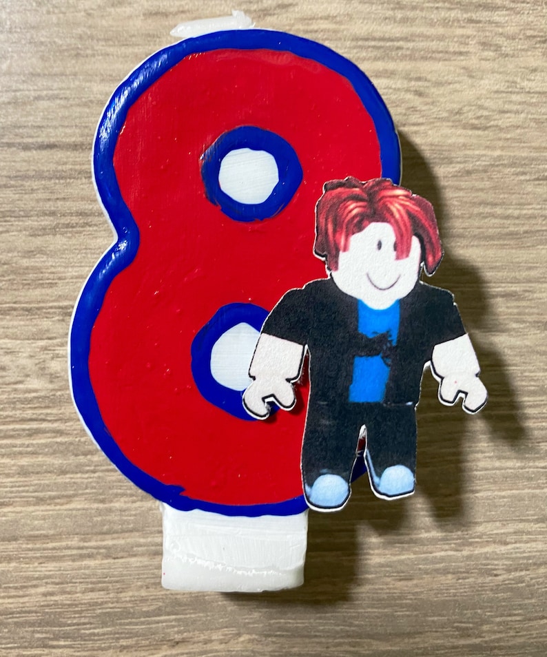Puede incluir: Vela de cumplea&ntilde;os n&uacute;mero ocho roja y azul con un personaje de Roblox de pie frente a ella. El personaje tiene el pelo rojo, una camisa negra, pantalones azules y una cara blanca.