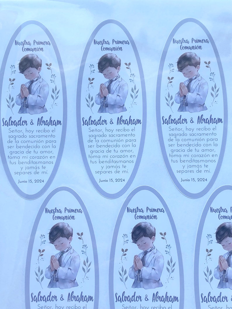 May include: Four oval-shaped stickers with a light blue background and a white border. Each sticker features a drawing of a young boy in a white suit with a light blue shirt, praying with his hands clasped. The boy is surrounded by light blue and green floral designs. The text "Nuestra Primera Comuni&oacute;n" is at the top of each sticker. The text "Salvador & Abraham" is at the bottom of each sticker. The text "Se&ntilde;or, hoy recibo el sagrado sacramento de la comuni&oacute;n para ser bendecido con la gracia de tu amor, toma mi coraz&oacute;n en tus benditasmanos y jam&aacute;s te separes de m&iacute;. Junio 15, 2024" is in the center of each sticker.