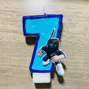 Puede incluir: Vela de cumplea&ntilde;os azul y blanca con forma de n&uacute;mero siete con un personaje de dibujos animados sosteniendo una espada en la parte delantera. El personaje lleva una m&aacute;scara negra y ropa negra y blanca.