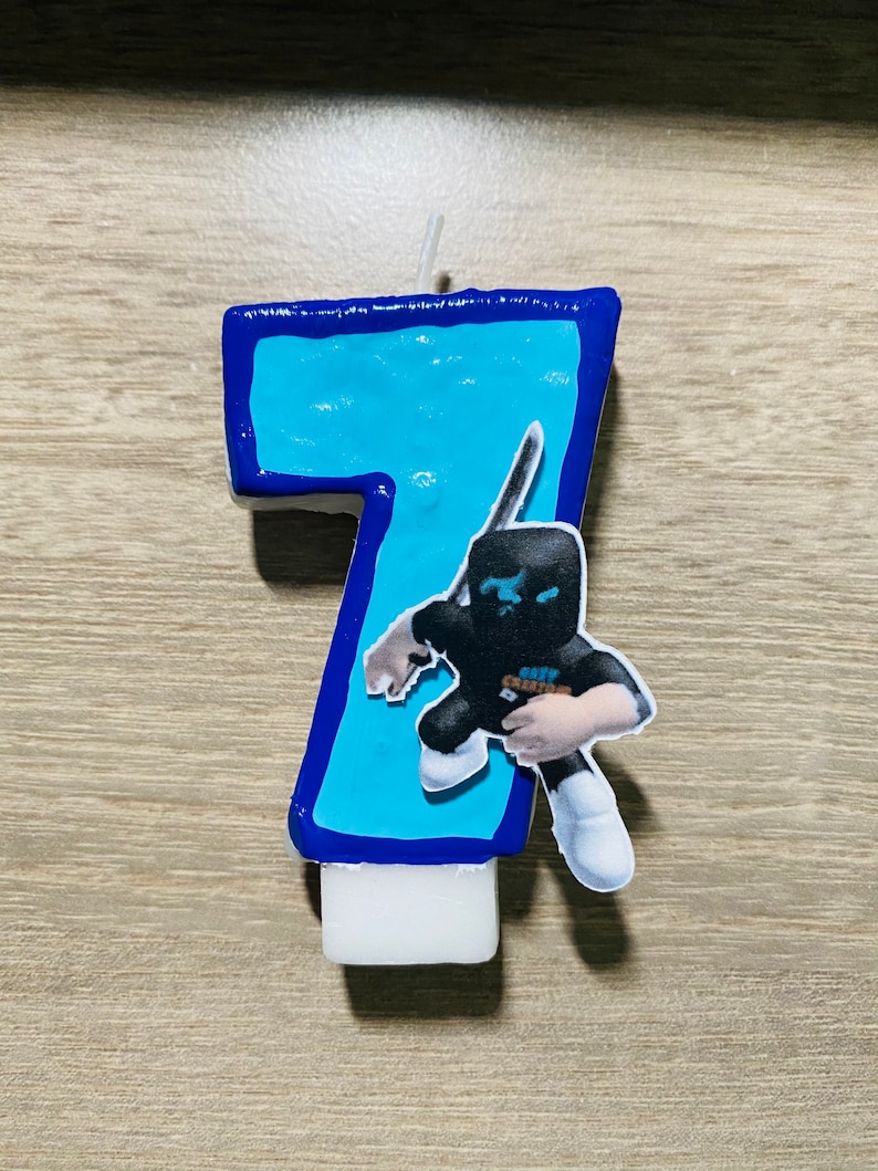 Puede incluir: Vela de cumplea&ntilde;os azul y blanca con forma de n&uacute;mero siete con un personaje de dibujos animados sosteniendo una espada en la parte delantera. El personaje lleva una m&aacute;scara negra y ropa negra y blanca.