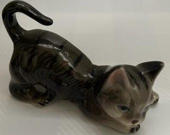 May include: A ceramic figurine of a gray tabby cat with black stripes, lying on its belly with its head turned to the side. The cat has a realistic appearance with detailed fur and facial features.