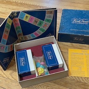 May include: A blue box with the game Trivial Pursuit inside. The box is open and the game board, cards, and playing pieces are visible. The game board is a blue circle with a track around the outside. The cards are in two stacks, one blue and one red. The playing pieces are small plastic figures.
