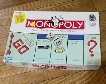May include: A Monopoly board game box with a red, white, and blue color scheme. The box features the iconic Monopoly logo and the text "Property Trading Game from Parker Brothers". The box also includes a picture of the game board with the "GO" space, the "Boardwalk" property, the "Luxury Tax" space, the "Park Place" property, and the "Chance" space. The box is designed for ages 8 to adult.