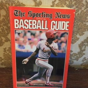 May include: The Sporting News Official Baseball Guide from 1986. The cover features a baseball player in a red hat and white uniform with the word "Cardinals" on the front. The player is holding a bat and is in a batting stance.