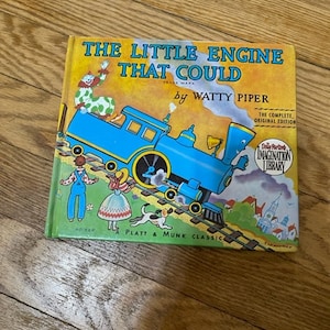 May include: A vintage children's book titled "The Little Engine That Could" by Watty Piper. The cover features a blue train engine pulling a train of colorful boxcars. The book is part of the Dolly Parton Imagination Library.