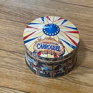 May include: A round tin box with a colorful design depicting a carousel with the text "Hersheypark Carousel Circa 1893". The top of the box features a red, white, and blue striped canopy with a blue star design.
