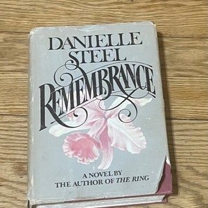 May include: A paperback book titled "Remembrance" by Danielle Steel. The cover features a pink and white orchid with a gray background and the text "A Novel by The Author of The Ring".