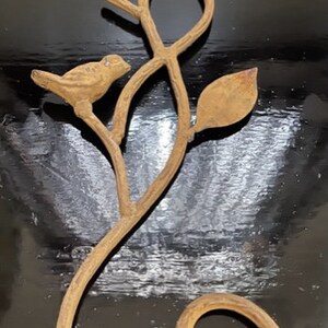 May include: A metal wall decor featuring a stylized branch with two birds perched on it. The branch is curled into a loop at the bottom, and the metal has a rustic, aged finish.