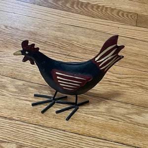 May include: A black metal chicken figurine with red and white stripes. The chicken has a long neck and a small head with a red comb. The chicken is standing on a wooden surface.