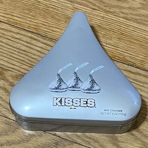 May include: A silver, triangular-shaped Hershey's Kisses tin. The tin features three images of Hershey's Kisses with their paper plumes. The word "KISSES" is printed in bold, black letters. The tin contains 4 oz (113 g) of milk chocolate.