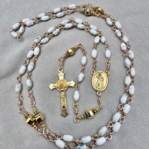 May include: A rosary featuring white marble-patterned beads linked by gold-toned wire. The piece includes a gold-toned crucifix with the letters INRI and a central oval medal depicting a figure with rays, connected by smooth, polished gold-toned beads.