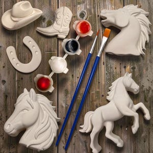 Pode incluir: Um conjunto de artesanato com figuras de cerâmica branca, incluindo duas cabeças de cavalo, um cavalo inteiro, um chapéu de cowboy, uma bota de cowboy e uma ferradura. Inclui quatro potes de tinta vermelha, preta, branca e vermelha, e dois pincéis azuis.
