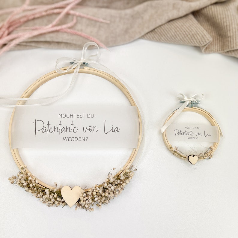 May include: Two wooden embroidery hoops with white ribbon and dried flowers. The larger hoop has a white card with the text "M&Ouml;CHTEST DU PATENTANTE VON Lia WERDEN?" The smaller hoop has a white card with the text "M&Ouml;CHTEST DU PATENTANTE VON Lia WERDEN?"