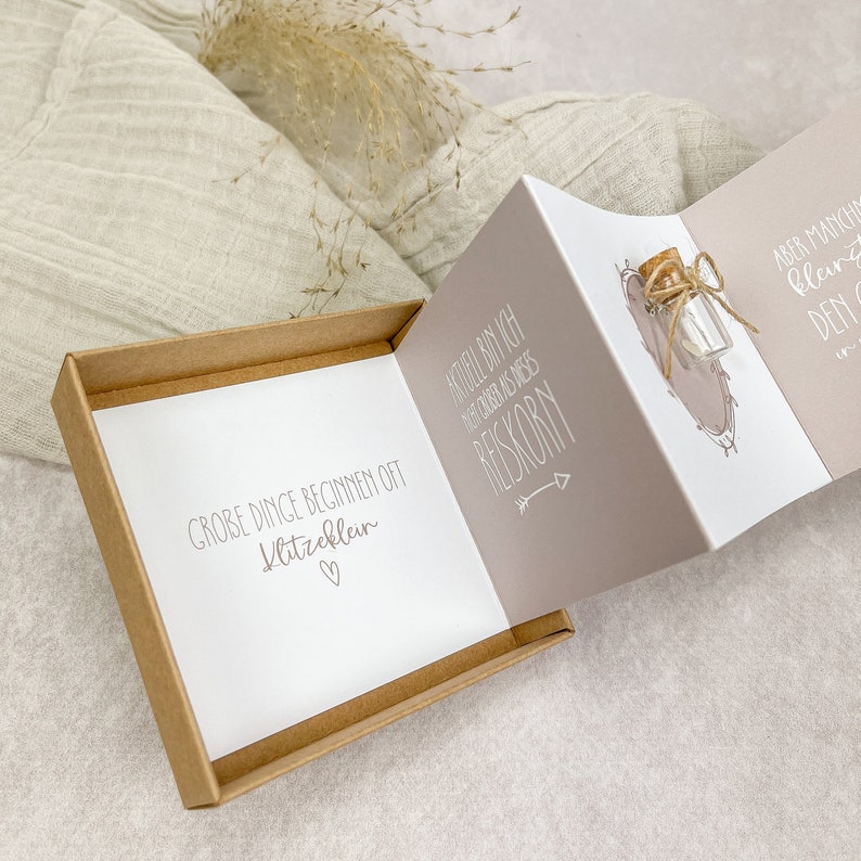 May include: A small, brown box with a white card inside. The card has the text "GROBE DINGE BEGINNEN OFT Klitzeklein" and a small heart. The box is open and a folded card is visible. The card has the text "AKTUEL BIN ICH NIGHT COOKER ANIS DIESES RESKORN" and an arrow pointing to the right. There is a small glass bottle with a cork stopper and twine tied around it.