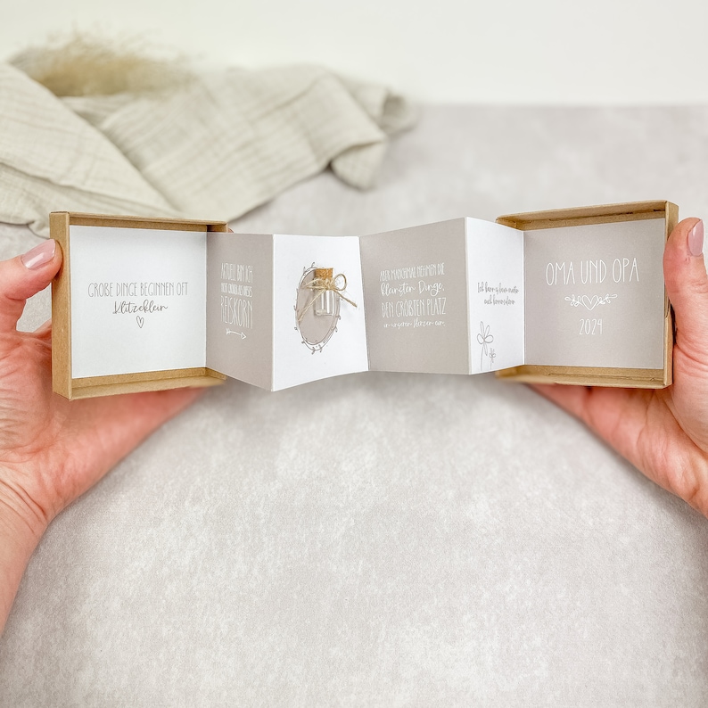 May include: A small, brown box with a white interior. The box is open and folded out to reveal a series of cards with text and illustrations. The text on the cards reads "Gro&szlig;e Dinge beginnen oft mit Kleinigkeiten", "Kleine Dinge machen das Leben sch&ouml;n", "Willkommen zum ersten Tag im neuen Jahr", "Bis hier hin und noch viel weiter", "Oma und Opa 2021".