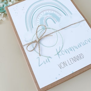 May include: A light blue and green watercolor rainbow with white clouds and gold glitter. The card is tied with twine and has the text "Zur Kommunion von Lennard".