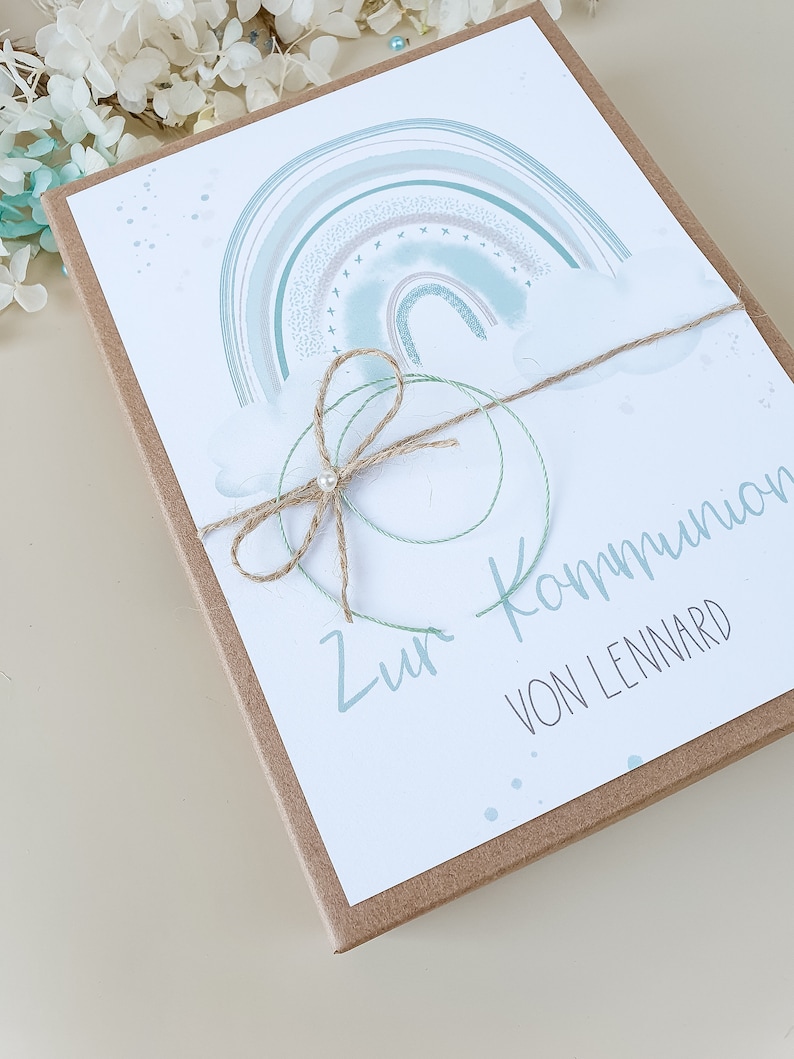May include: A light blue and green watercolor rainbow with white clouds and gold glitter. The card is tied with twine and has the text "Zur Kommunion von Lennard".